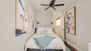 Une chambre à coucher avec un lit, un ventilateur de plafond, des œuvres d’art accrochées aux murs et une porte donnant sur une autre pièce.