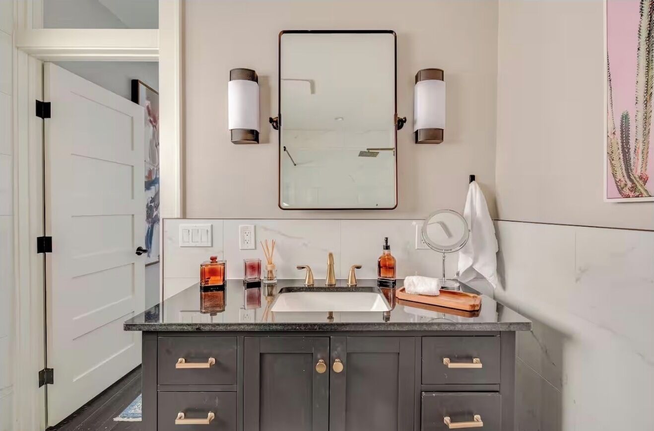 Une salle de bain avec un grand miroir, deux appliques murales, un lavabo avec un plan de travail et un porte-serviettes.