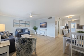 A modern living room with a sofa, armchair, and coffee table, a dining area with a table and chairs, and a kitchen with cabinets and appliances. - Gulf Coast Beach Life - Treasure Island (Treasure Island)