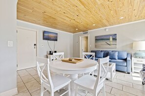 Una habitación con techo de madera, una mesa redonda blanca con sillas, un sofá azul y un televisor montado en la pared.