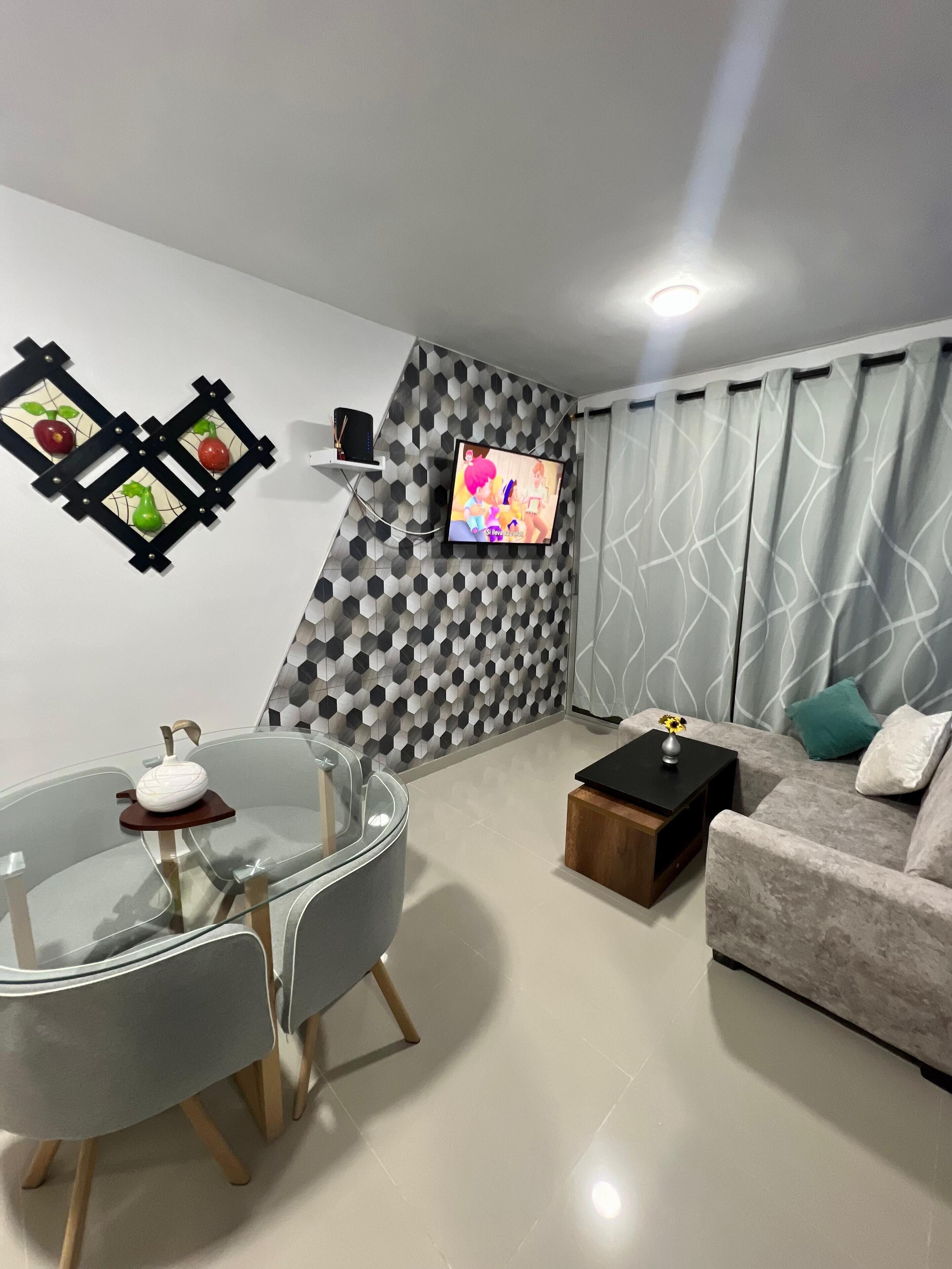 Een moderne woonkamer met een glazen eettafel, een bank, een televisie bevestigd aan een muur met een geometrisch patroon, en een klein houten bijzettafeltje.