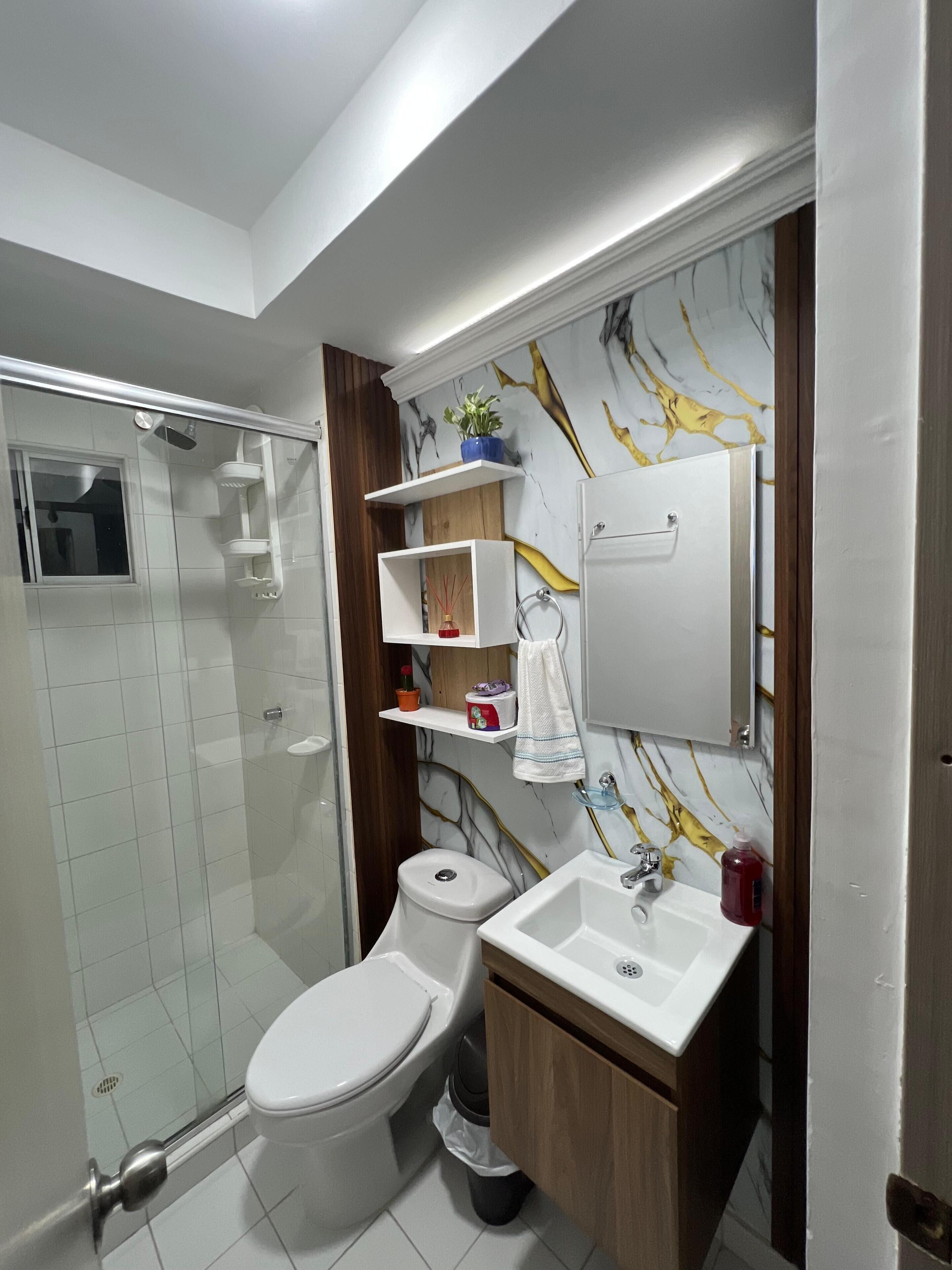 Une salle de bain moderne avec une douche à parois de verre, des toilettes blanches, un lavabo avec un miroir au-dessus et un mur à motifs de marbre.