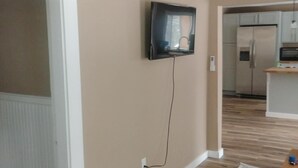 A room with a television mounted on the wall, a wooden floor, and a kitchen area in the background.