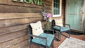 Une véranda en bois avec une pancarte indiquant « MAISON », deux chaises avec des coussins, un vase de fleurs et un tapis à motifs.