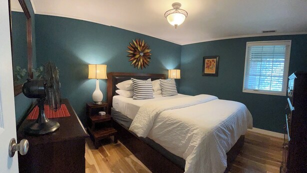 Une chambre à coucher comprenant un lit, deux lampes de chevet, une horloge murale, un élément décoratif mural et une fenêtre avec des stores.