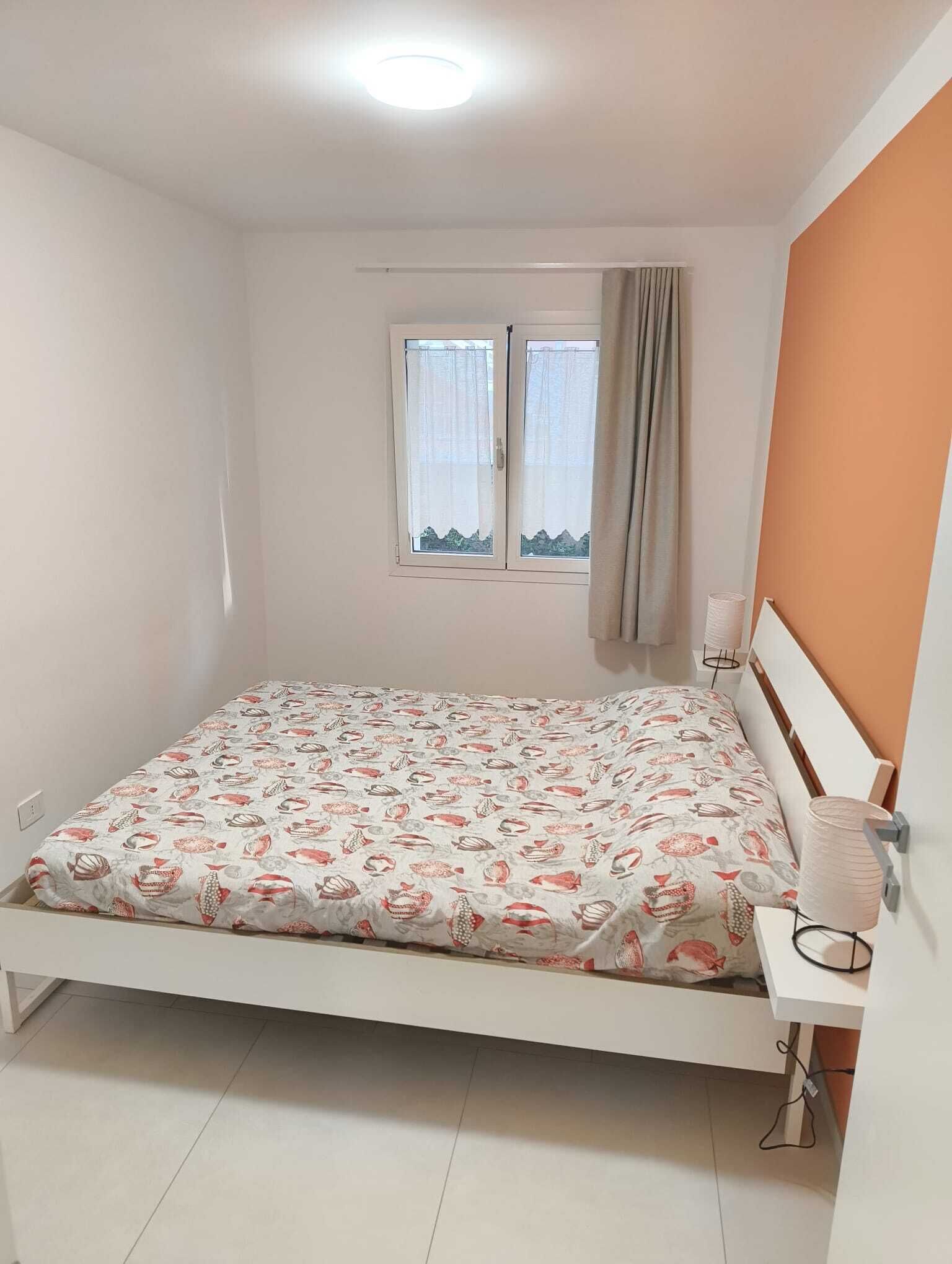 A small, single-bed room with a white bedspread featuring a fish pattern, a window with curtains, and a white headboard.