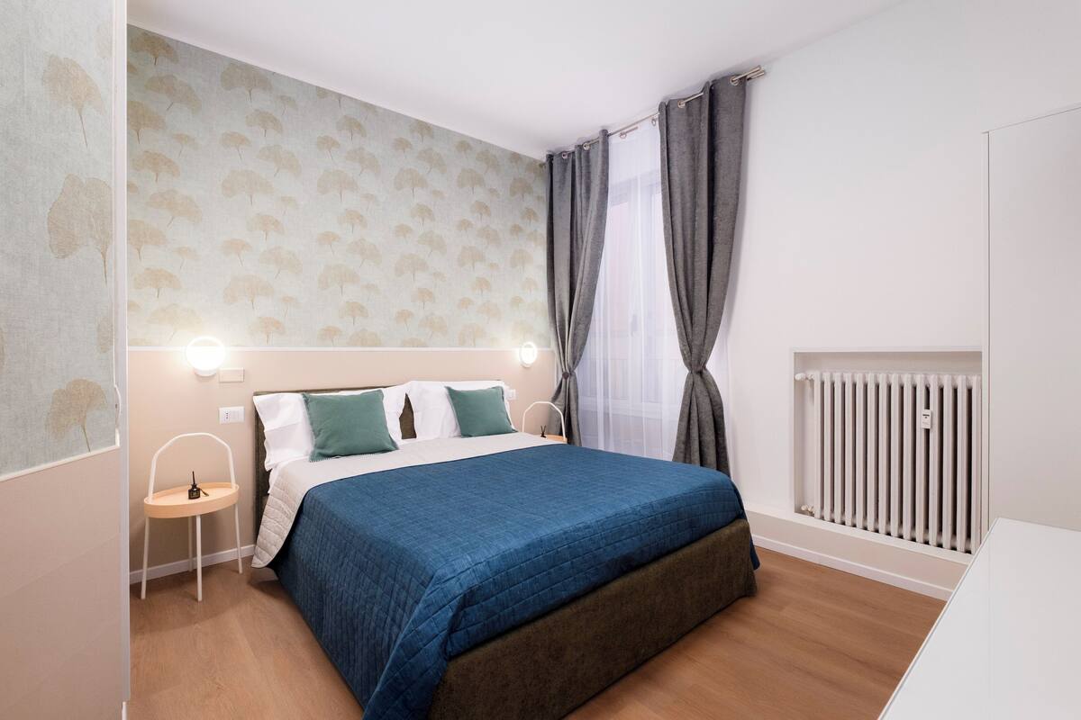 Une chambre à coucher comprenant un lit, une table de chevet, une lampe, des rideaux et un radiateur.