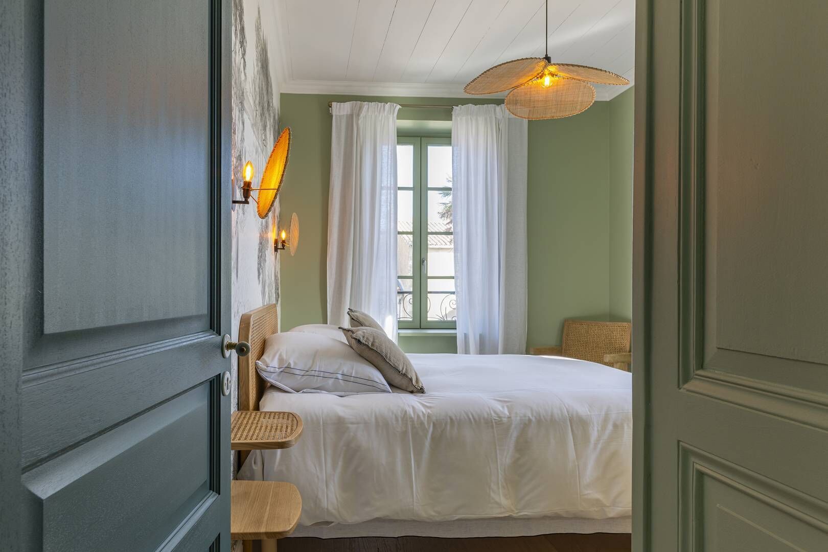 A bedroom with a bed, a window with curtains, a wall-mounted lamp, and a wicker chair.