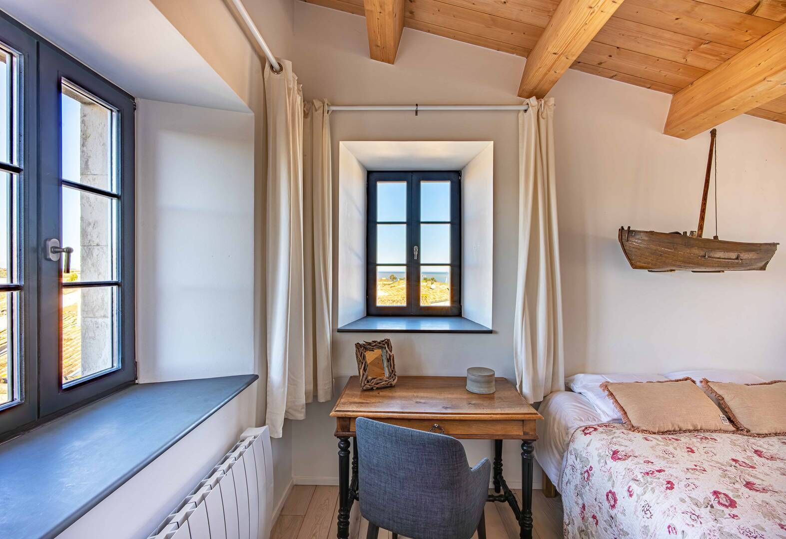 Un dormitorio con ventana, una mesa de madera, una silla y una cama con ropa de cama floral.