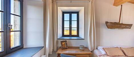 Un dormitorio con ventana, una mesa de madera, una silla y una cama con ropa de cama floral.