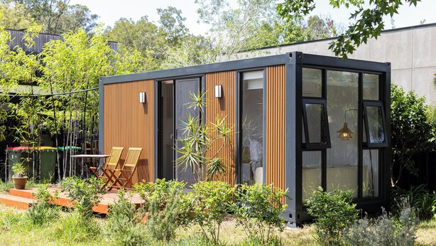 A modern, compact house with a wooden exterior, large windows, and a patio area with outdoor furniture.