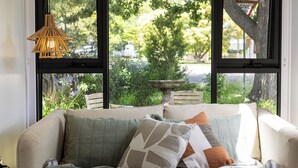 A cozy room with a large window, a wicker pendant light, a cushioned sofa with multiple pillows, and a view of greenery outside.