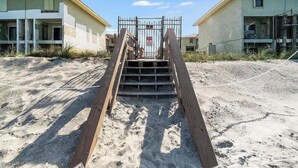 Propiedad frente a la playa con entrada privada, escalones de madera que conducen a un portón metálico y dos edificios con ventanas y balcones a la vista.