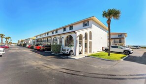 Um hotel com vários andares, fachada branca, várias varandas e um estacionamento com diversos carros.
