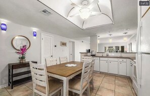 Uma área de refeições com mesa de madeira e cadeiras brancas, uma cozinha com armários brancos e um ventilador de teto.