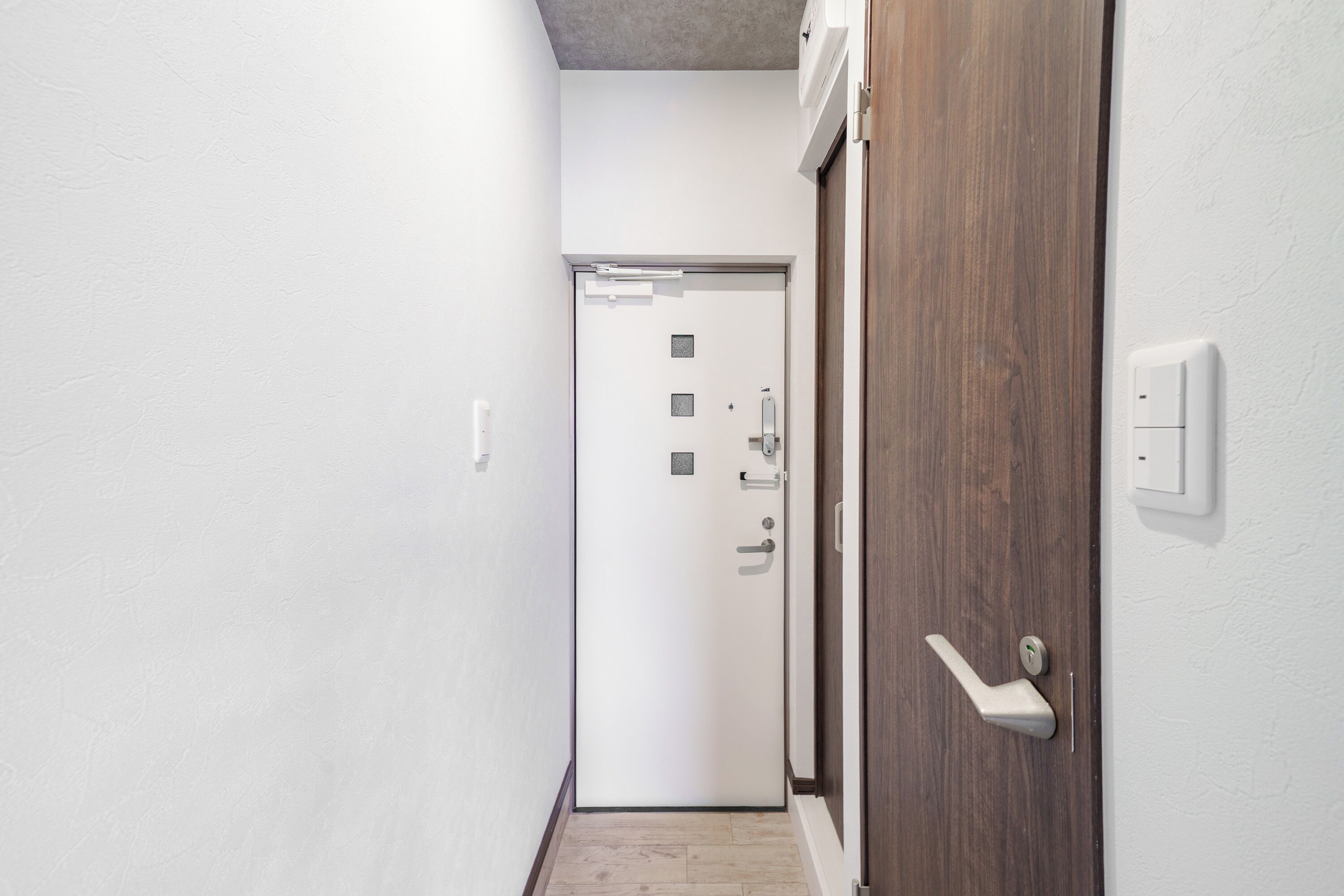 A narrow hallway with a white door at the end, a wooden door on the right, and a light switch on the wall.
