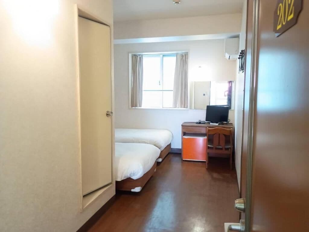A hotel room with a bed, a desk, a chair, a window with curtains, and a door with a number.