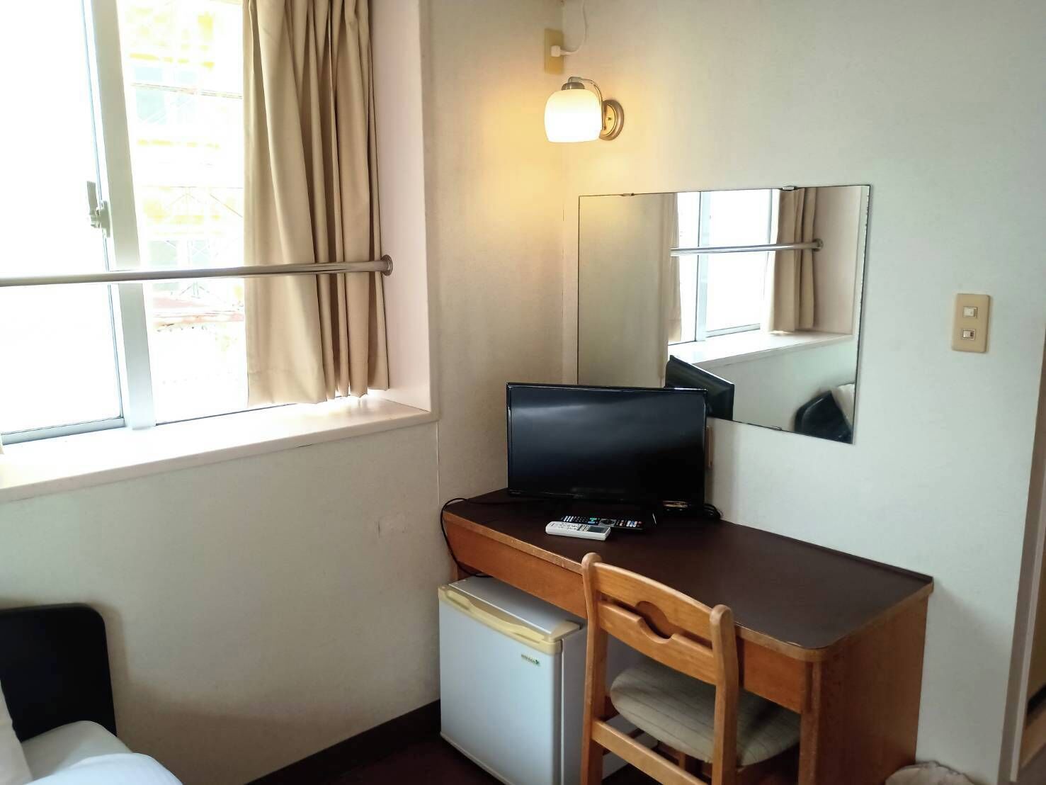 A small room with a desk, chair, and a refrigerator. There is a window with curtains and a wall-mounted light fixture.