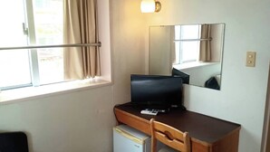 A small room with a desk, chair, and a refrigerator. There is a window with curtains and a wall-mounted light fixture. - HotelSumi (Naha)