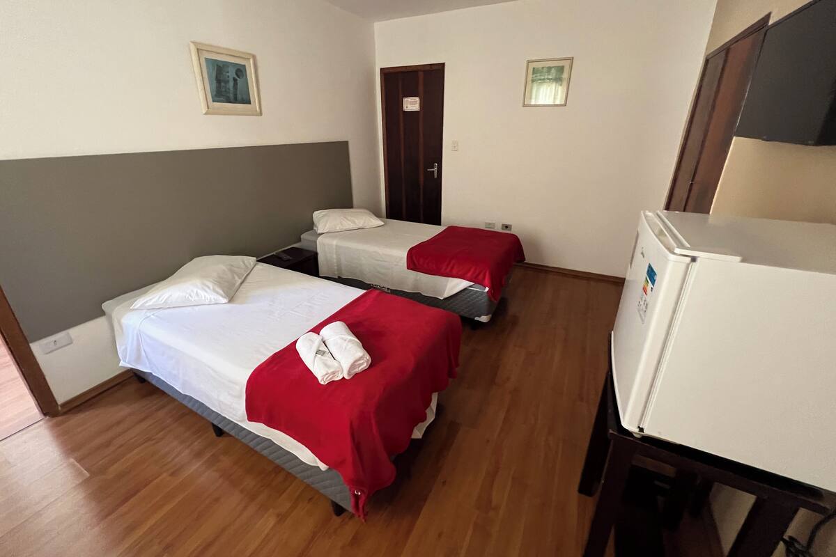 Quarto de hotel com duas camas individuais, piso de madeira, geladeira branca e porta com uma placa.
