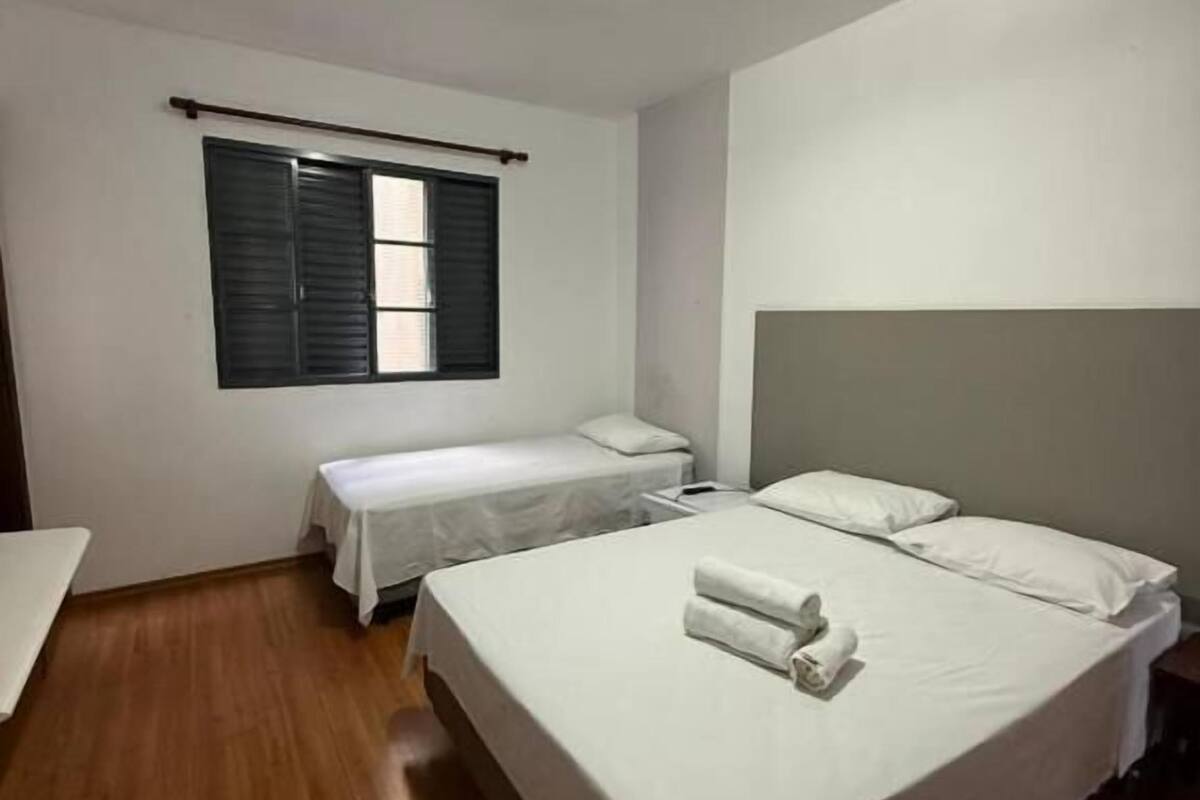 Quarto de hotel com duas camas de solteiro, uma pequena mesa, uma janela com persianas e uma luminária no teto.