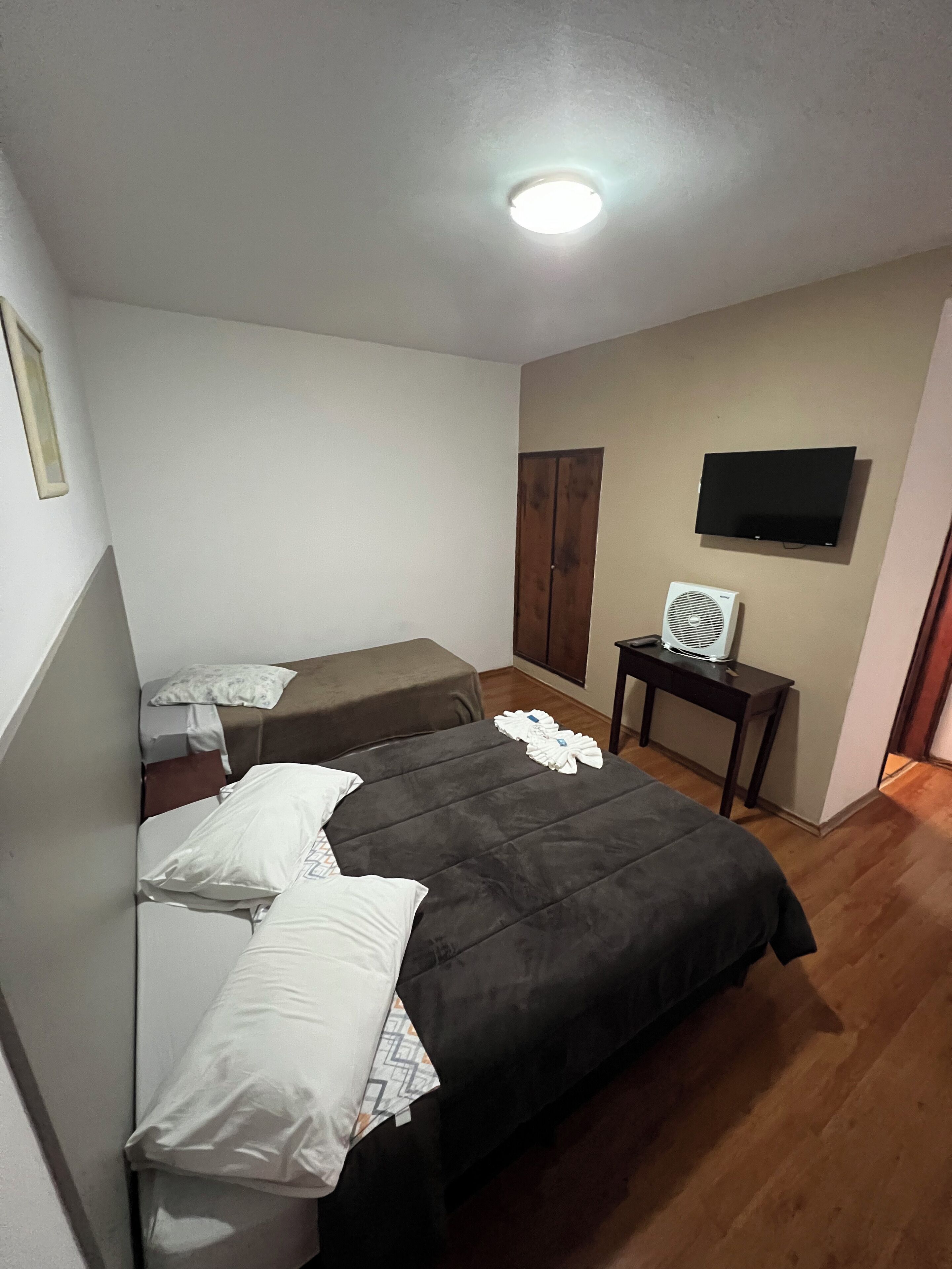 A hotel room with two beds, a TV, a desk with a fan, and a wooden floor.