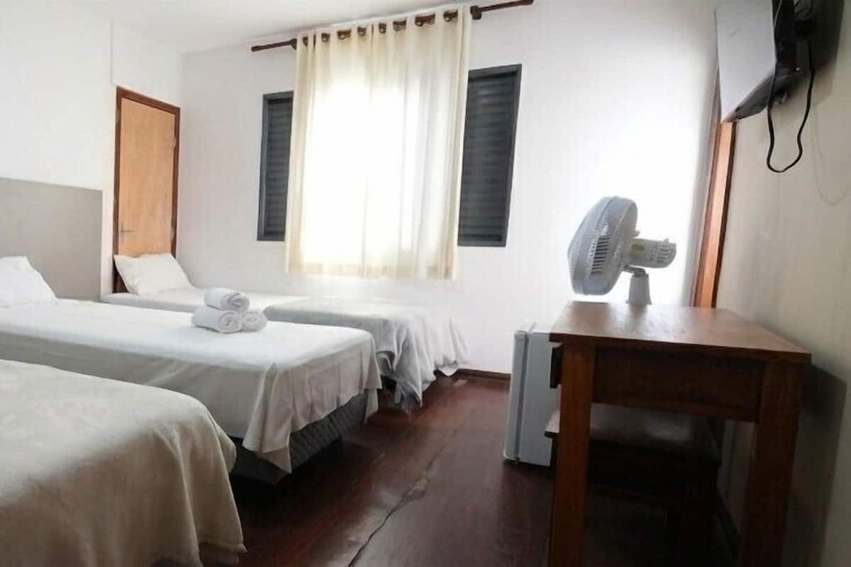 Quarto de hotel com duas camas de solteiro, uma escrivaninha de madeira, uma cadeira, um ventilador e uma janela com cortinas.