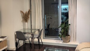 A modern living room with a glass coffee table, a dining area with a glass table and chairs, a large window with curtains, and a potted plant. - Luxury Canary Wharf 1 bed apt (London)