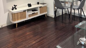 A modern living room with a flat-screen TV mounted on the wall, a glass dining table with two chairs, a white console table with wicker baskets, and a vase with dried plants. - Luxury Canary Wharf 1 bed apt (London)