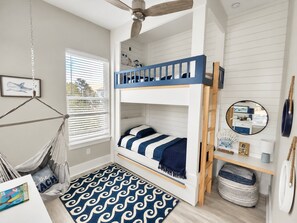 Una cama litera con baranda azul, una silla colgante, un espejo redondo y un estante de madera con varios objetos.