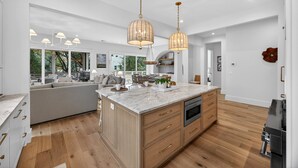 A modern kitchen with a large island, wooden cabinets, and a built-in oven. There are pendant lights above the island and a dining area with a sofa and table. - BRAND NEW BUILD; Gorgeous home with POOL, Hot Tub, and steps to BEACH! (Isle Of Palms)