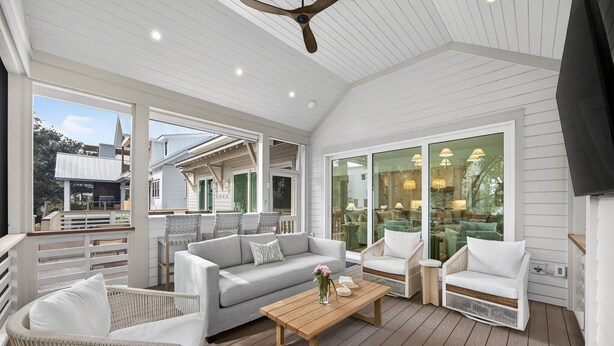 A covered outdoor seating area with a wooden deck, white furniture, and large glass doors leading to an indoor space. - BRAND NEW BUILD; Gorgeous home with POOL, Hot Tub, and steps to BEACH! (Isle Of Palms)