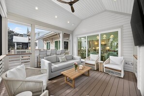 Uma área de estar coberta ao ar livre, com deck de madeira, móveis brancos e grandes portas de vidro que dão para um espaço interno.