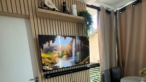 Un salon moderne avec un téléviseur à écran plat fixé sur un mur recouvert de panneaux en bois, un canapé en cuir noir, une petite table ronde avec un vase, et une porte sur le côté gauche.