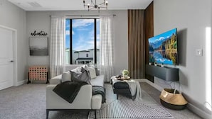 Ein modernes Wohnzimmer mit einer Couch, einem Couchtisch, einem Fernseher und einem großen Fenster mit Vorhängen.