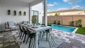 Eine moderne Terrasse mit Esstisch für vier Personen, Deckenventilator und Blick auf einen Pool im Garten.