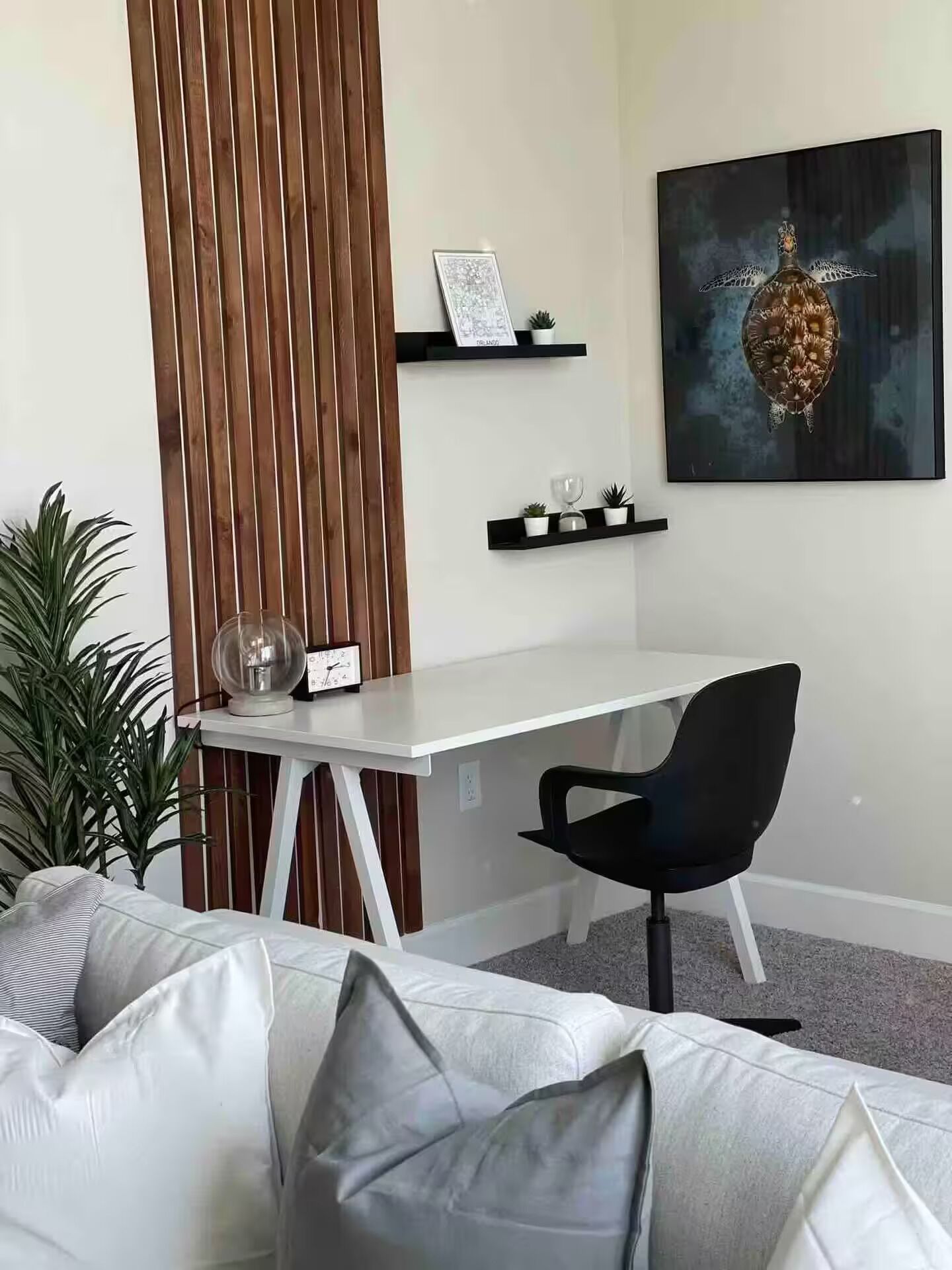 A modern room with a white desk, black chair, and a white sofa with grey cushions. There are two black shelves on the wall, one with a framed picture and the other with small plants and a glass object. A large abstract painting is on the wall above the desk.