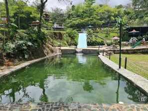 A tranquil outdoor pool area with a central waterfall, surrounded by lush greenery and a small bridge. - Aldea Los Pioneros (Venecia)