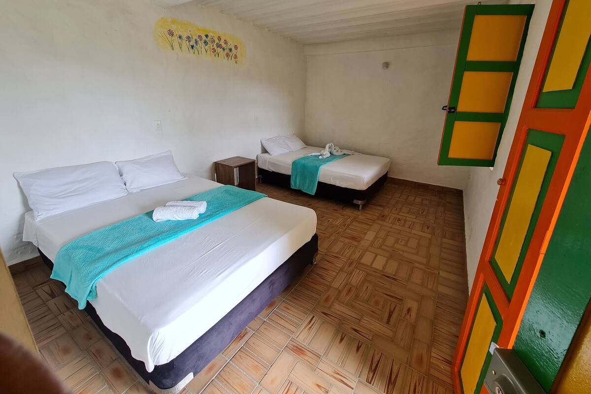 A room with two beds, each with white bedding and a teal blanket. There is a small wooden nightstand between the beds. The walls are plain, and the floor has a patterned tile design.