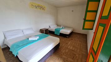 A room with two beds, each with white bedding and a teal blanket. There is a small wooden nightstand between the beds. The walls are plain, and the floor has a patterned tile design.
