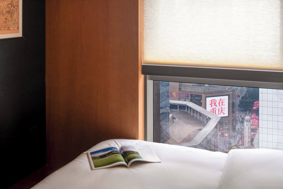 Ein Hotelzimmer mit Bett, Fenster mit Blick auf die Stadt und einer Zeitschrift auf dem Bett.