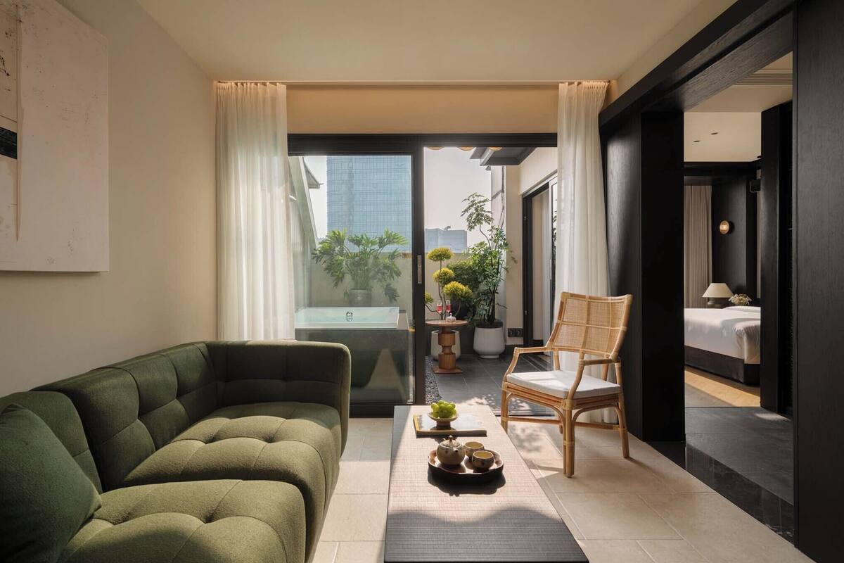 Ein modernes Wohnzimmer mit einem grünen Sofa, einem Holzstuhl und einem kleinen Tisch mit einer Ablage. Eine Schiebetür öffnet sich zu einem Balkon mit Pflanzen und Blick auf Gebäude.