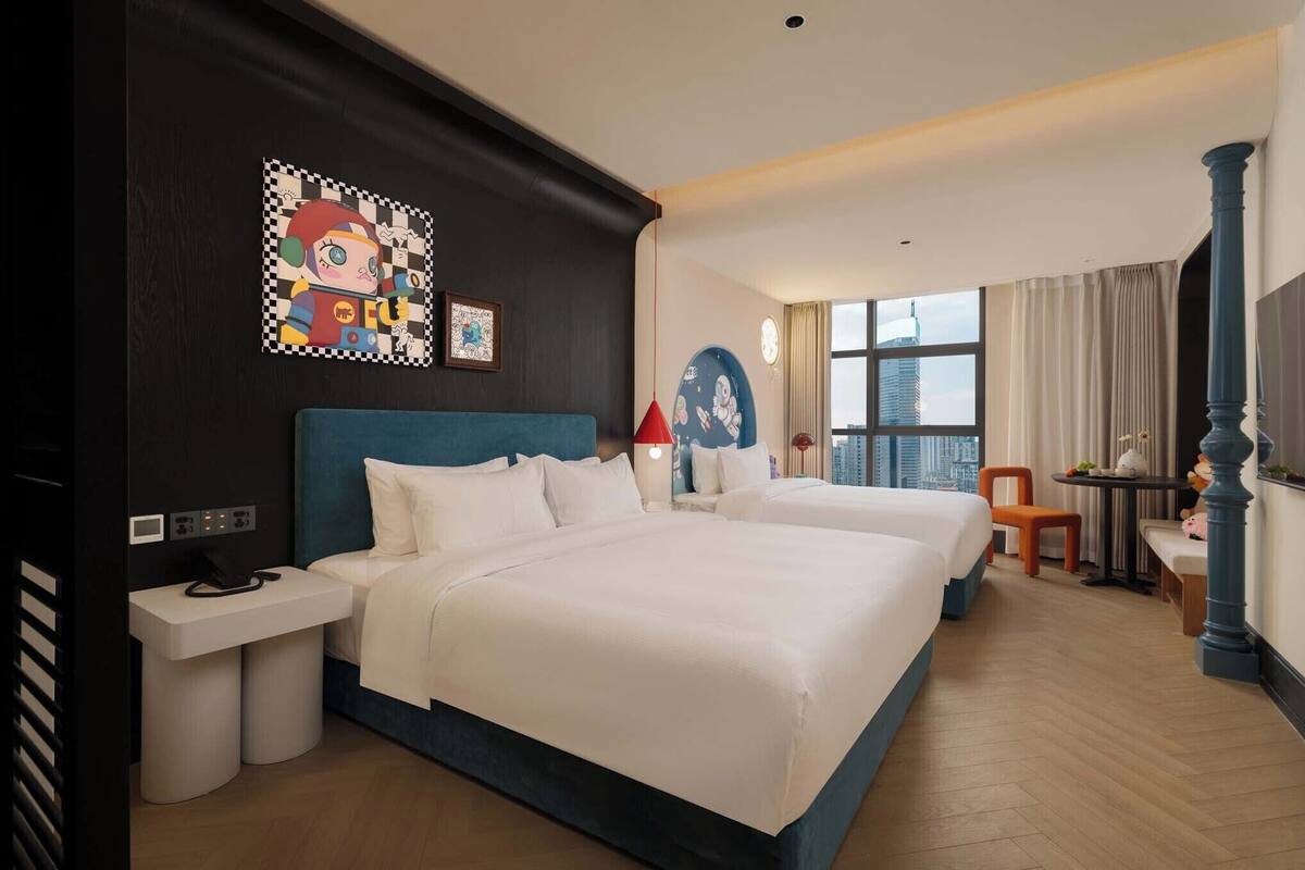 Ein Hotelzimmer mit zwei Betten, einem Schreibtisch, einem Sessel und Blick auf die Stadtlandschaft.