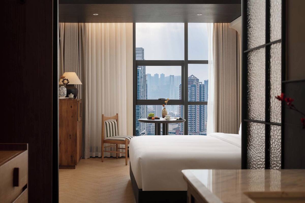 Ein Hotelzimmer mit großem Fenster, einem Bett, einem Sessel, einem kleinen Tisch mit Lampe und Blick auf hohe Gebäude.
