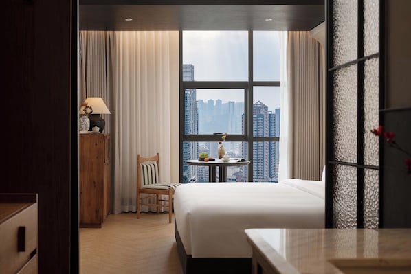Une chambre d’hôtel avec une grande fenêtre, un lit, une chaise, une petite table avec une lampe et une vue sur des immeubles de grande hauteur.