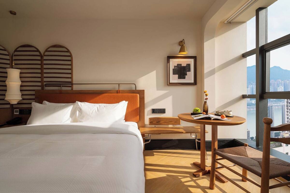 Ein Hotelzimmer mit einem großen Bett, einem Holznachttisch, einem kleinen runden Tisch mit Stuhl und Blick auf die Stadt durch ein großes Fenster.