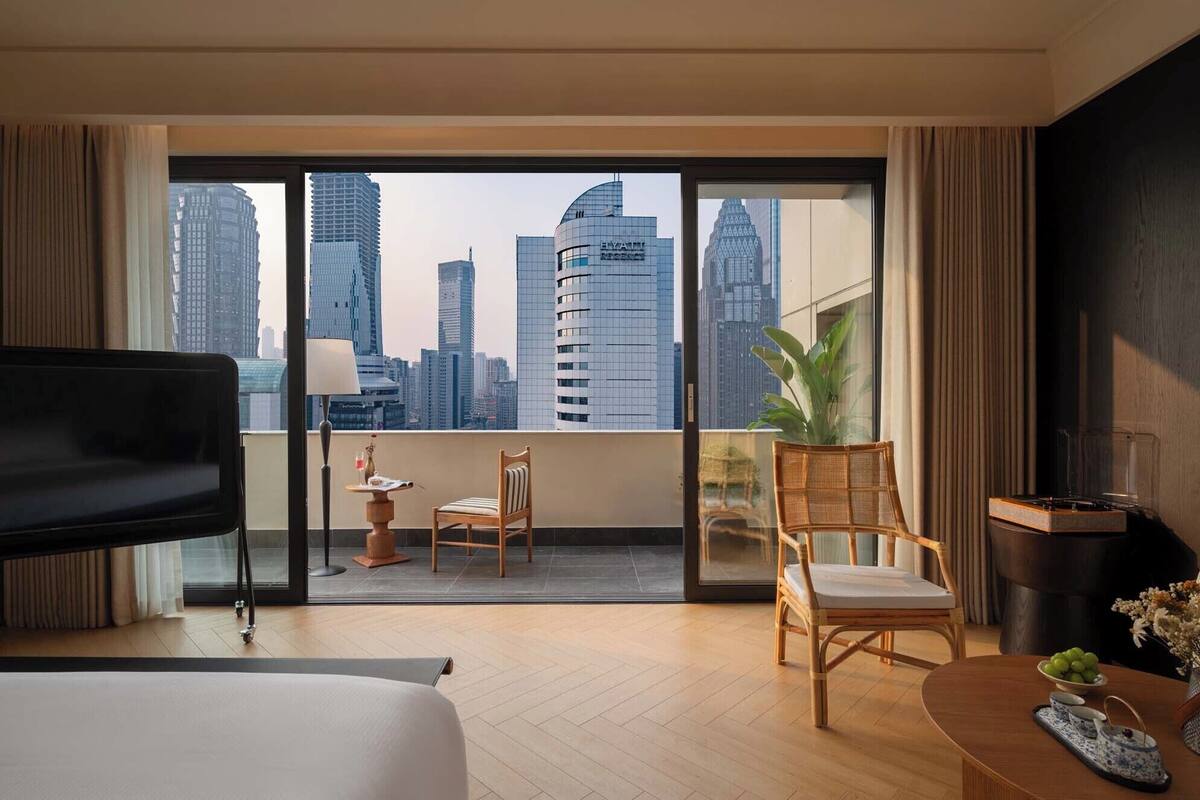 Ein modernes Hotelzimmer mit einem großen Balkon, einem Bett, einem Sessel, einem kleinen Tisch und Blick auf die Stadtlandschaft.