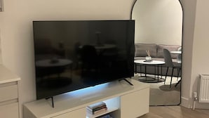 Ett modernt vardagsrum med en platt-TV på ett vitt TV-bord, en rund spegel och en matplats med ett bord och stolar.