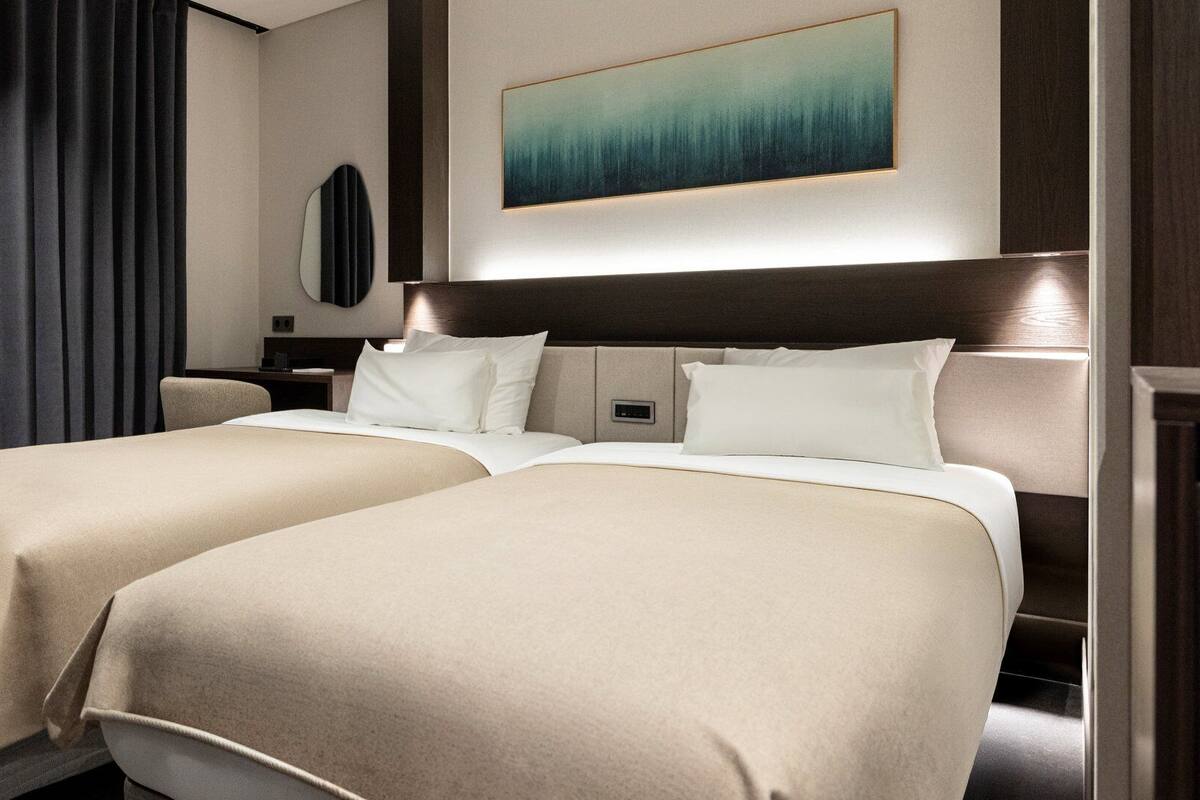 A hotel room with two beds, a desk, a chair, a painting on the wall, and a window with curtains.