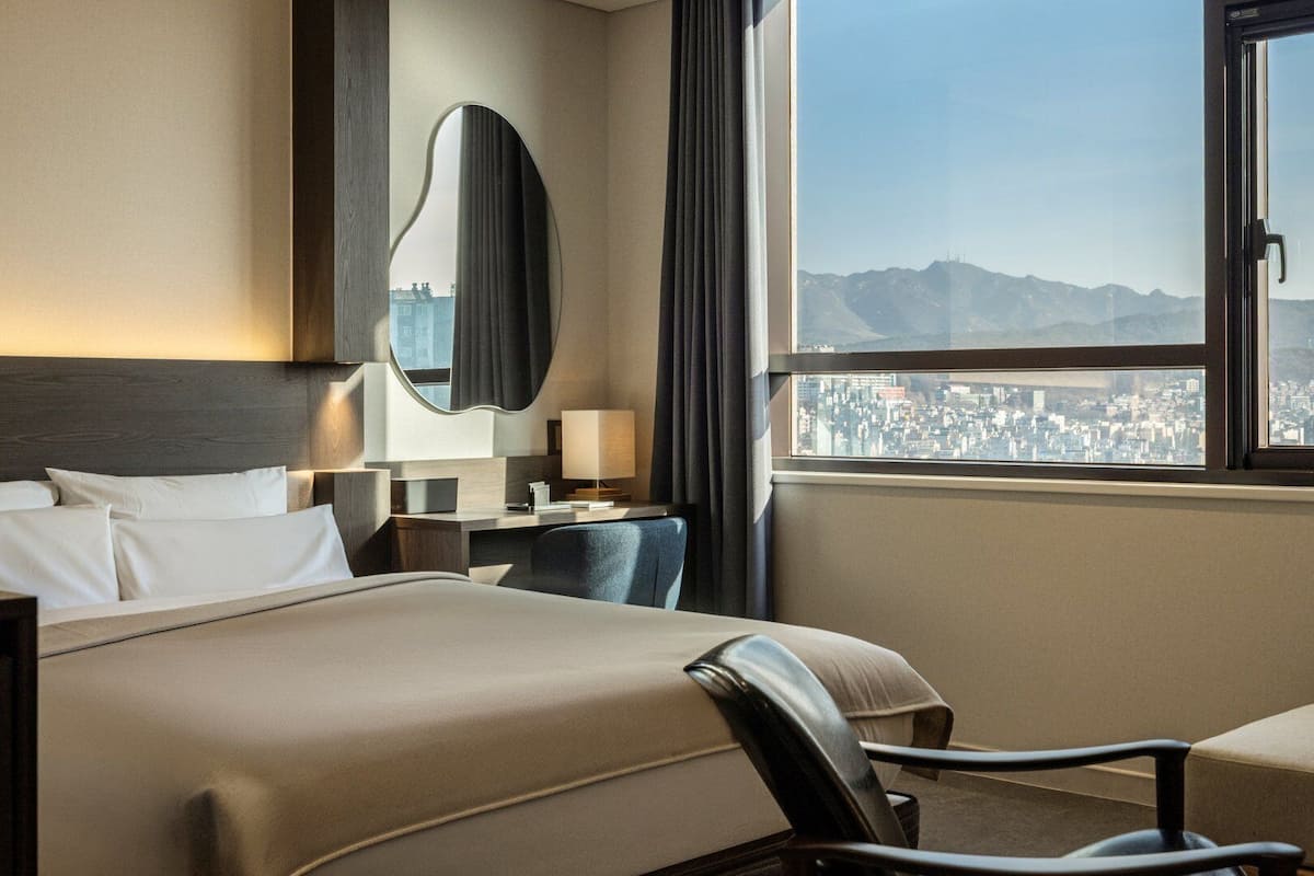 A hotel room with a bed, a desk, a chair, a mirror, and a window with a city view.
